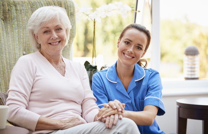 Portrait Of Senior Woman Sitting In Chair With Nurse In Retirement Home