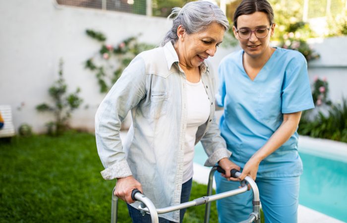 Young female nurse supporting old woman to walk outdoors with mobility walker, in retirement home