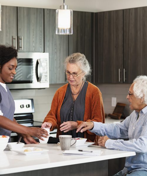 A home healthcare worker, an African-American woman in her 30s, helping a senior couple in their 80s in the kitchen. They are eating a healthy snack of fruit.